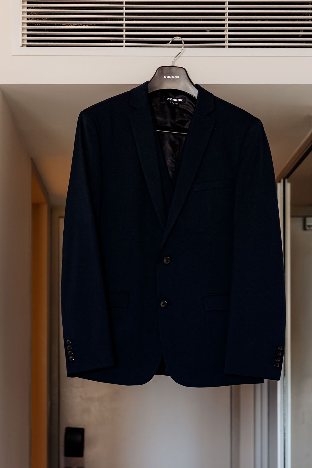 A sleek navy blue blazer jacket hangs on a clothing hanger against a white wall.