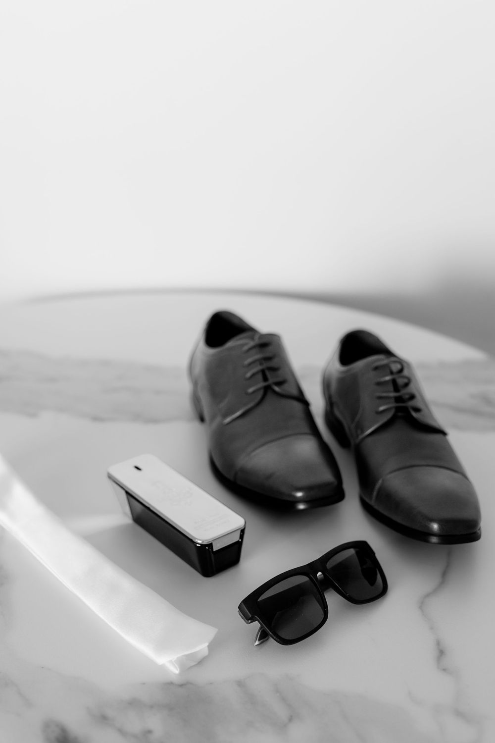 Black dress shoes, sunglasses, and mobile phone arranged on white marble surface in minimalist style.