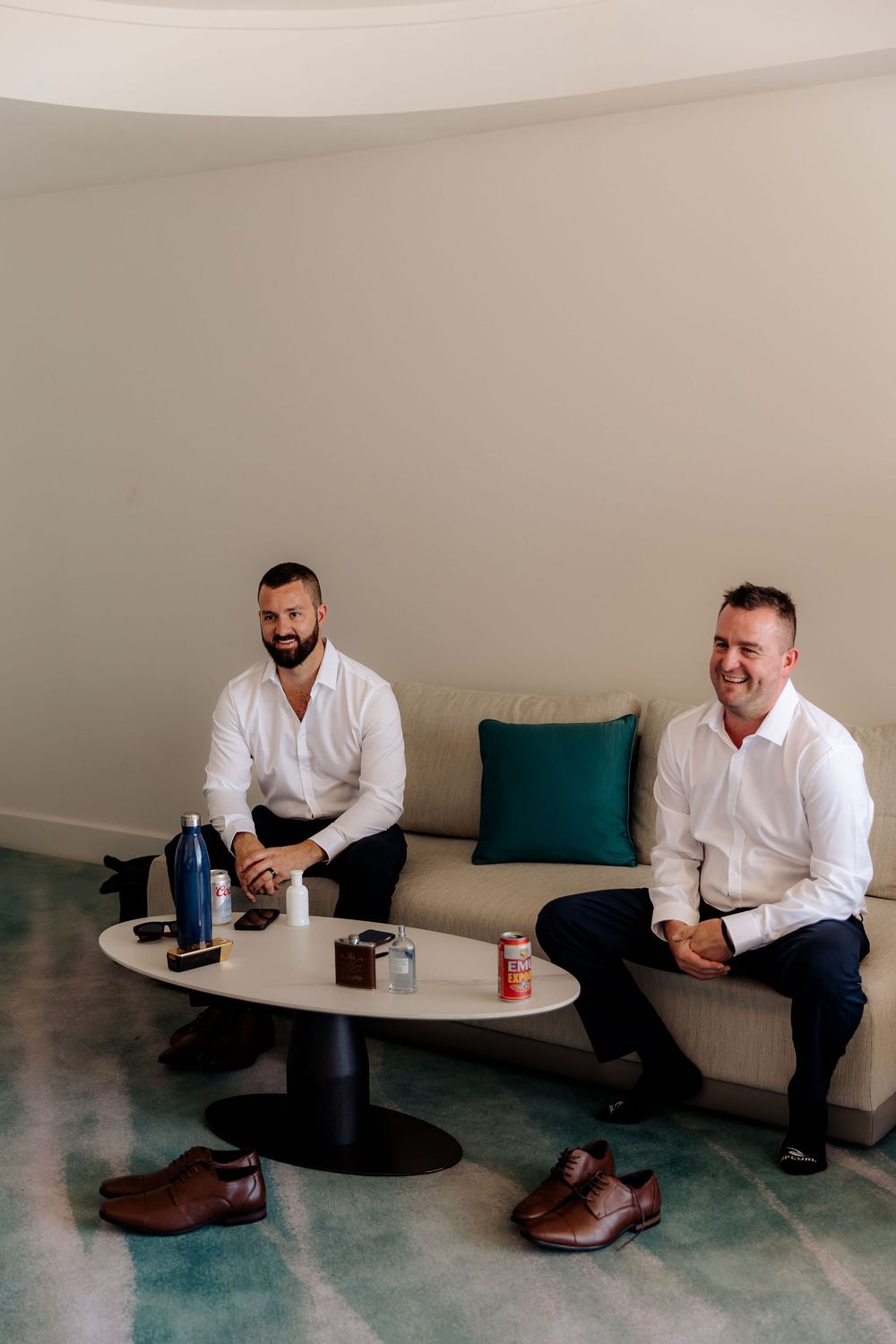 Two men in white shirts sitting on a bench with drinks and brown dress shoes placed on the floor in a minimal setting.