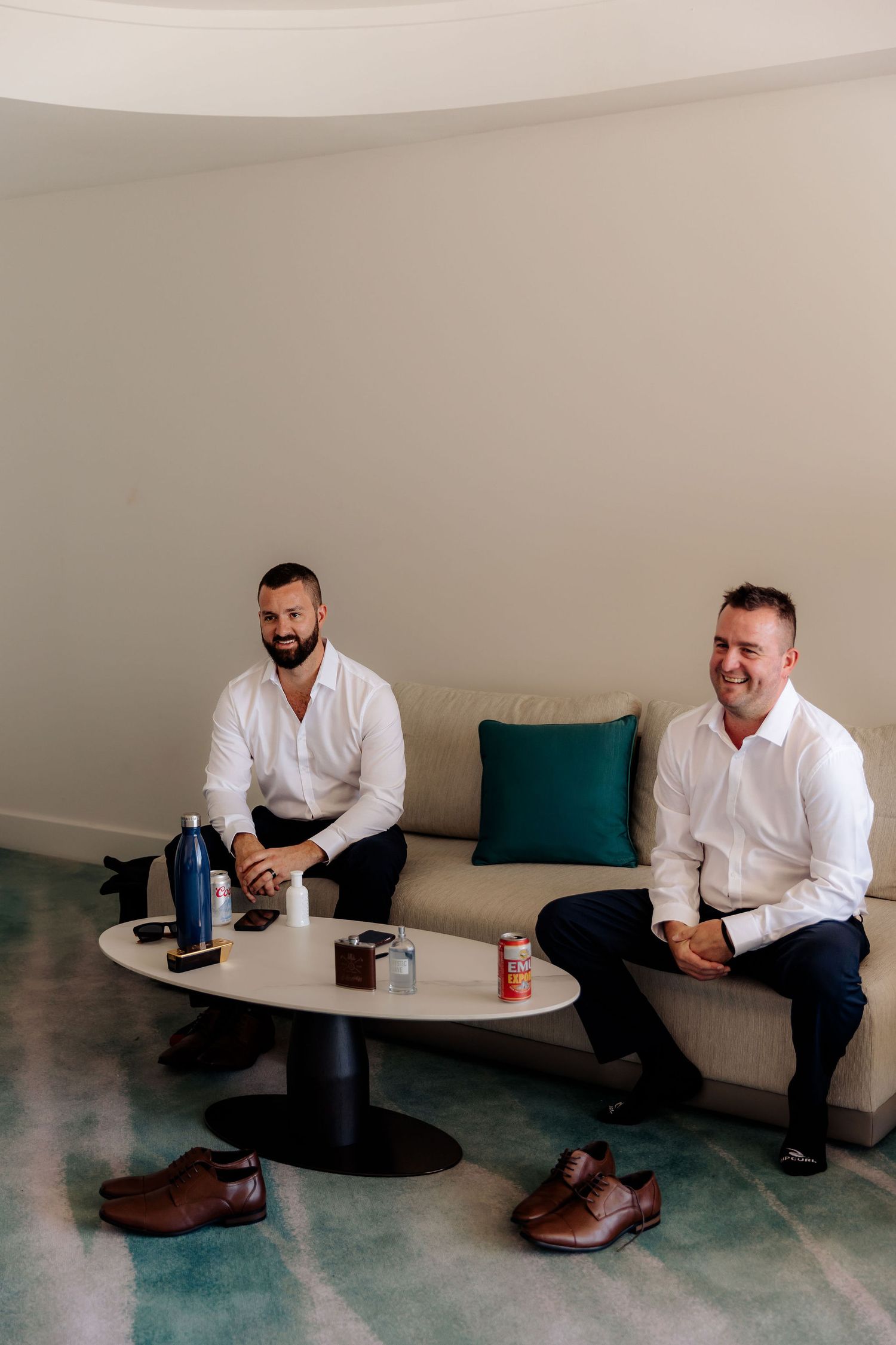 Two men in white shirts sitting on a bench with drinks and brown dress shoes placed on the floor in a minimal setting.