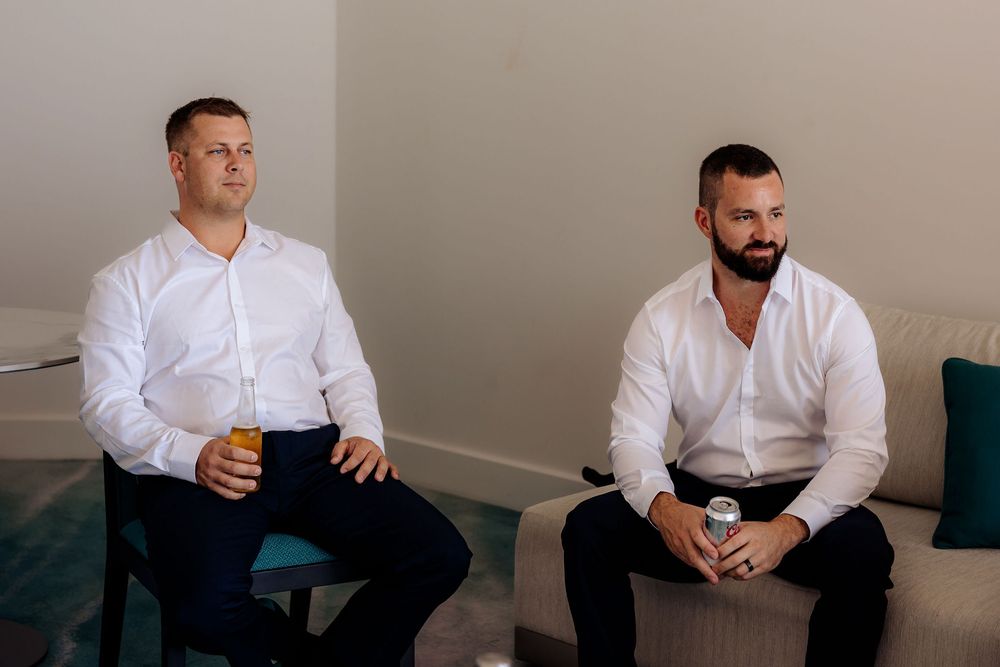 Two men wearing white shirts sit casually on a couch in an indoor setting.