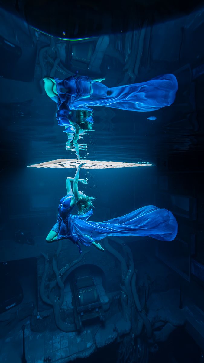 Ethereal underwater dancer in flowing blue dress creates mirror reflection effect in deep blue aquatic environment.
