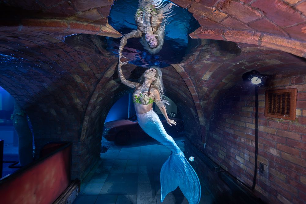 Dancer in blue dress performing graceful movements in dimly lit brick room with dramatic lighting effects.