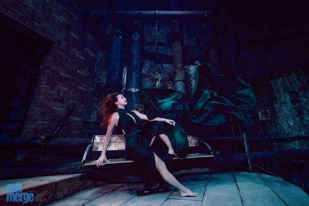 A person in black attire reclines against a vintage car in a dimly lit brick-walled setting with dramatic lighting.