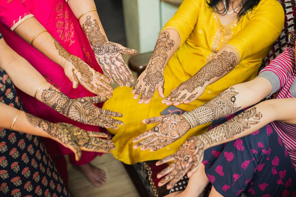 Multiple hands adorned with intricate henna designs are shown against colorful traditional Indian clothing.