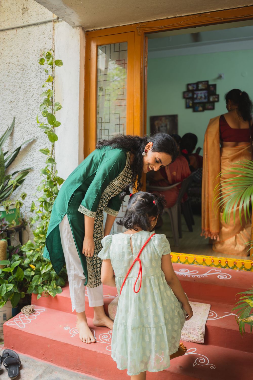 A woman in green bends down to greet a small child on steps decorated with rangoli patterns.