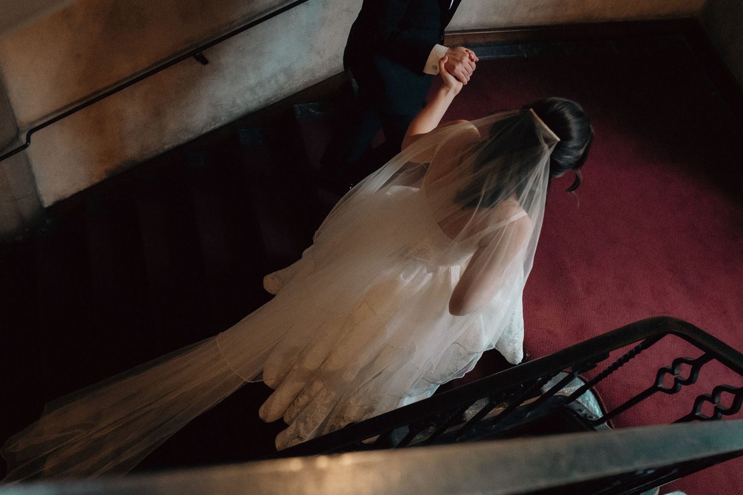 Bride in elegant white gown with long veil descending ornate staircase with red carpet.