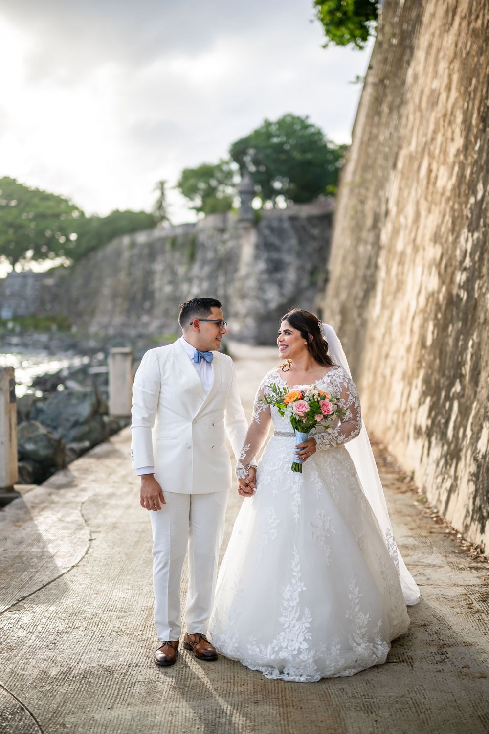 A romantic wedding photo series showing the couple posed along a historic stone wall in white attire with natural lighting.