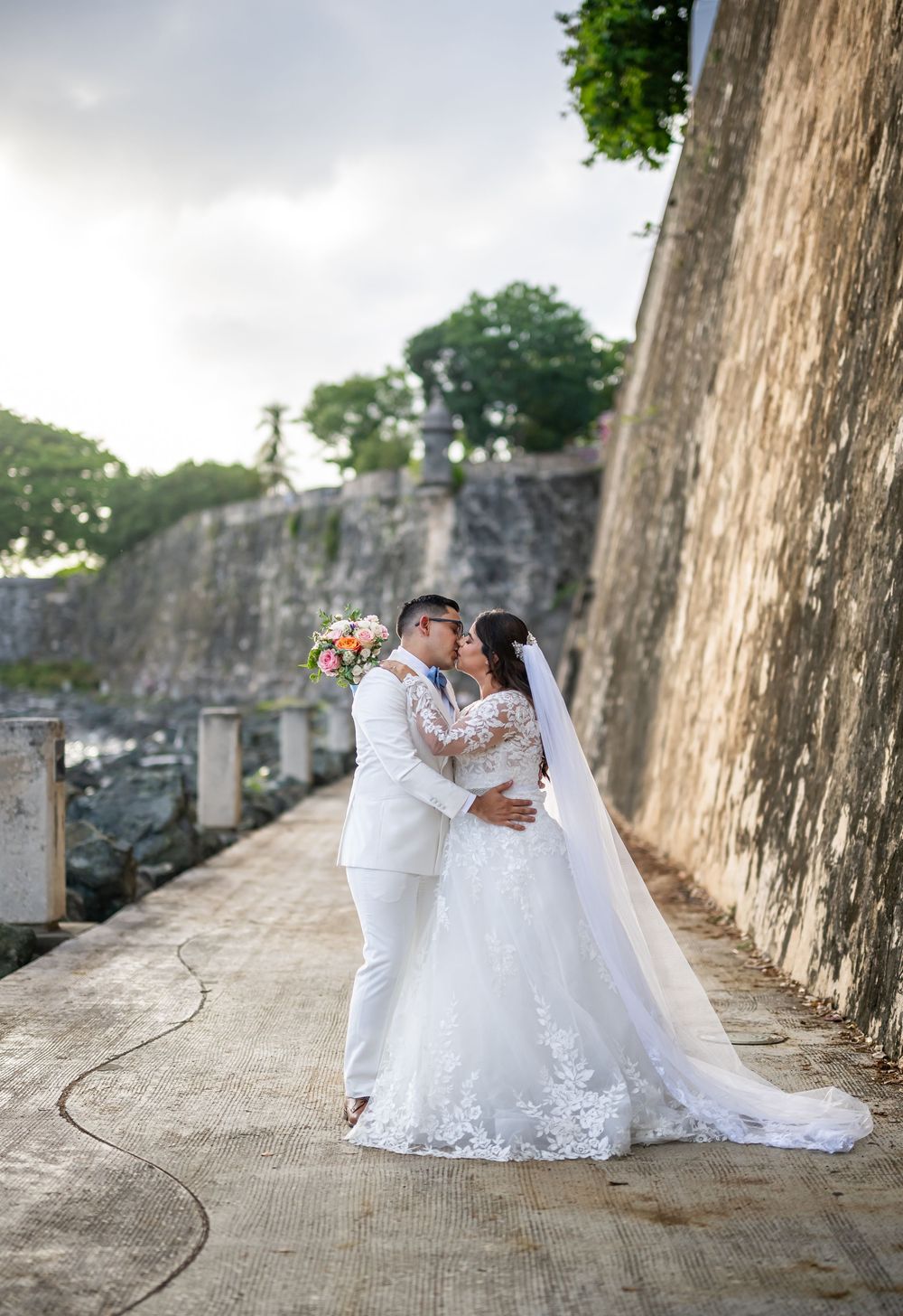 A couple in white wedding attire embrace by a stone wall in Old San Juan, Puerto Rico during an outdoor photo session.