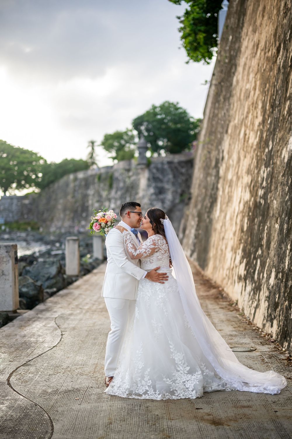 Wedding couple shares intimate moments in white attire against historic stone wall backdrop for artistic photo series.