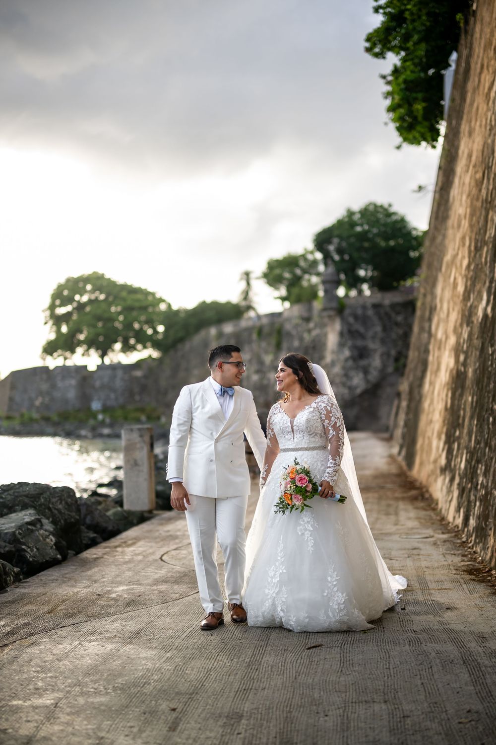 A wedding couple walks along a seaside path by stone walls on an overcast day.