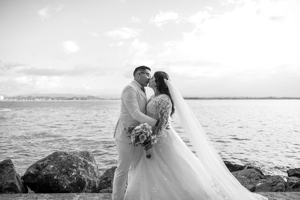 A romantic black and white wedding photo captured on a rocky shore with the ocean and cloudy sky in the background.