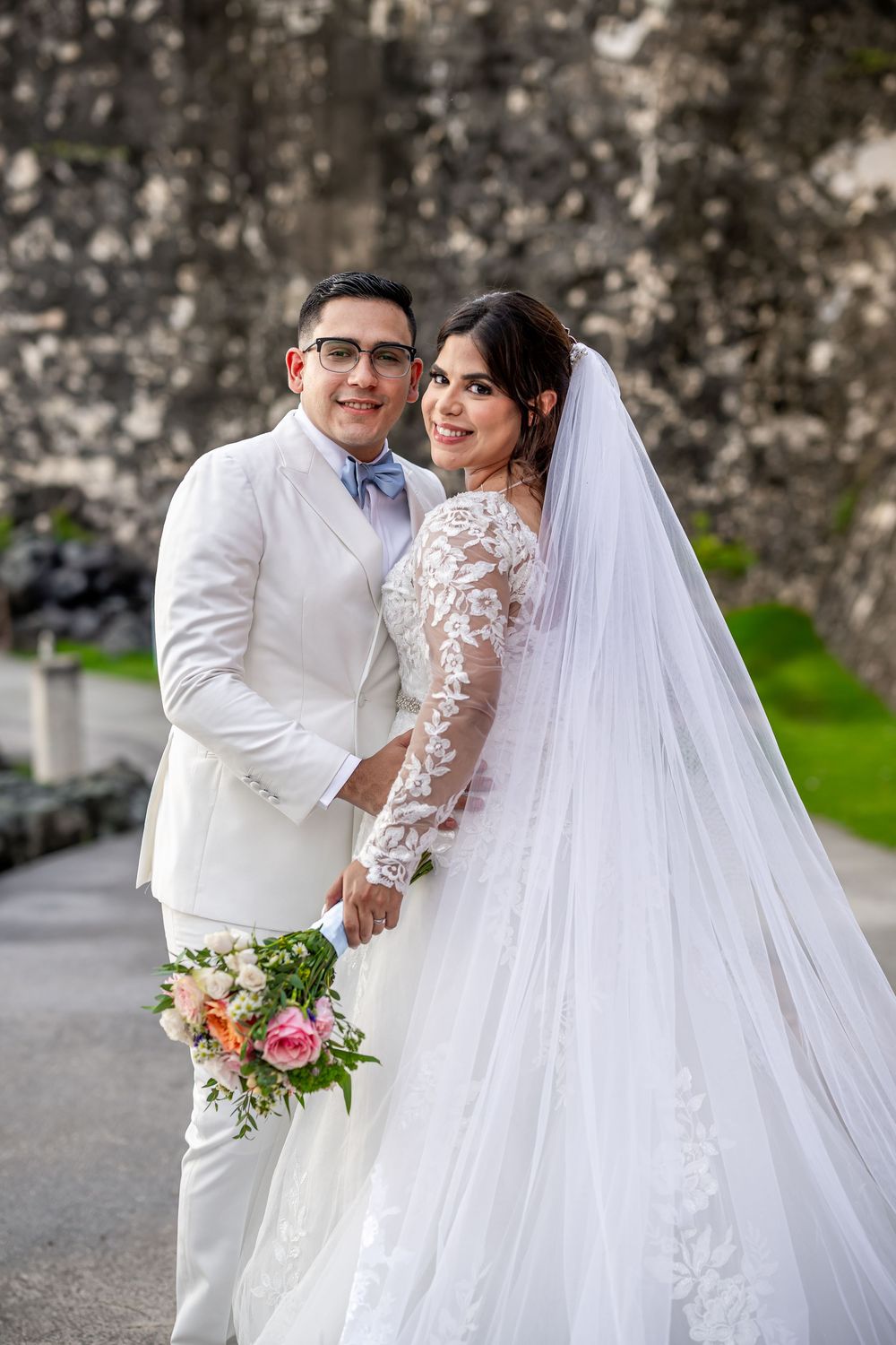 A newlywed couple poses outdoors in white attire, with the bride wearing a long-sleeved lace gown and veil holding a floral bouquet.