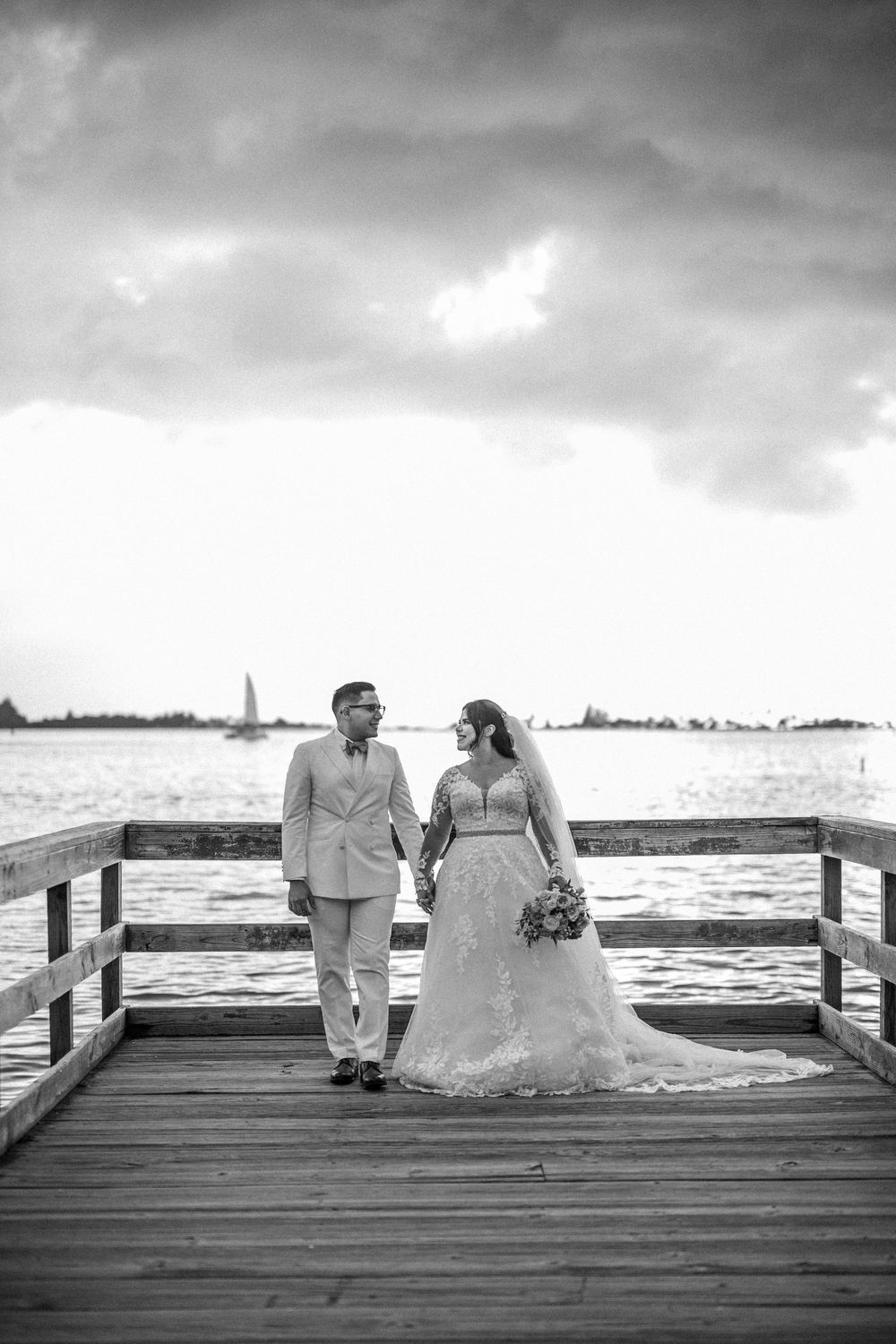 A couple sits together on a wooden pier overlooking the water on their wedding day in black and white.