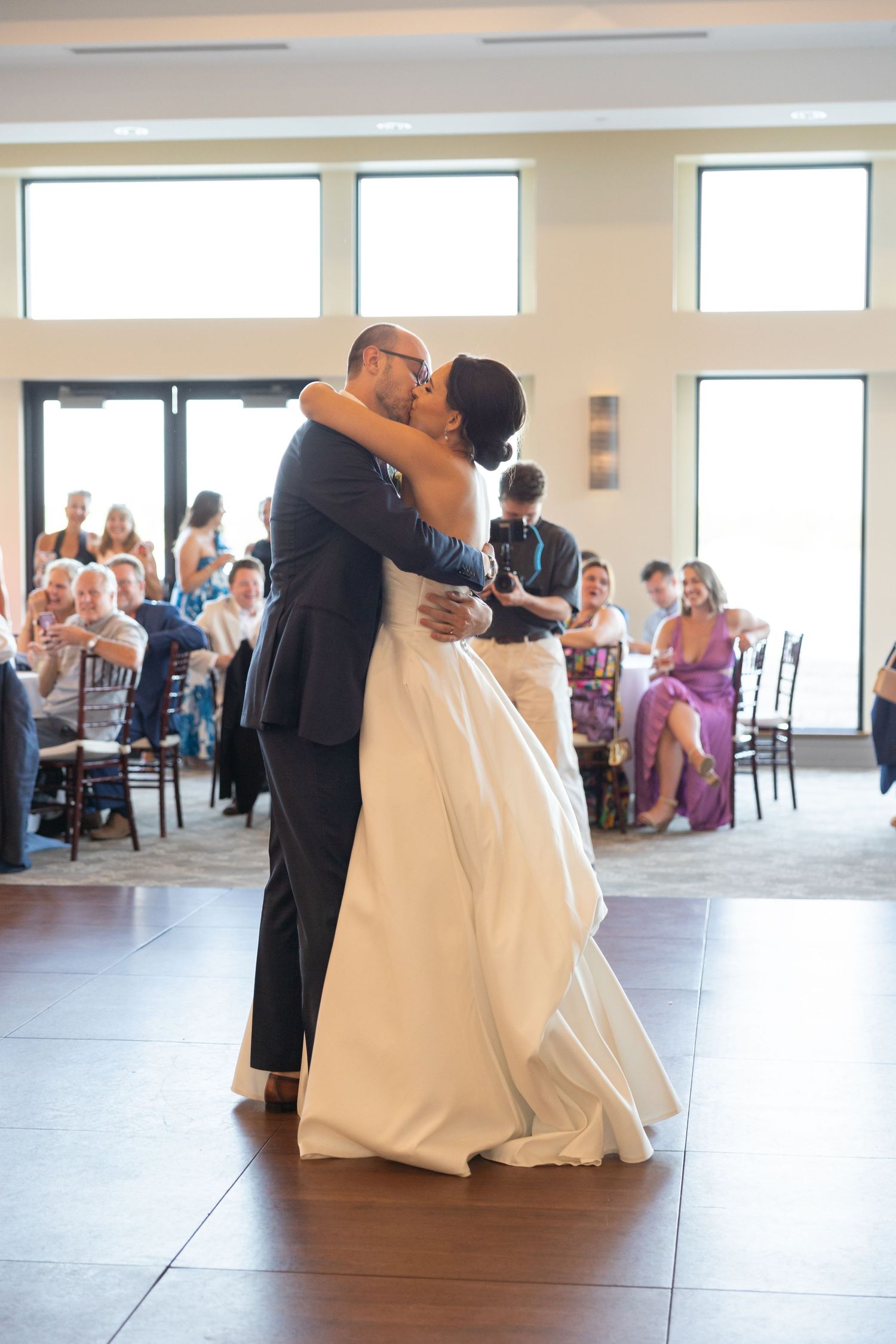 A couple shares their first dance at a wedding reception in a bright, modern venue with large windows and watching guests. Rachel Martinez