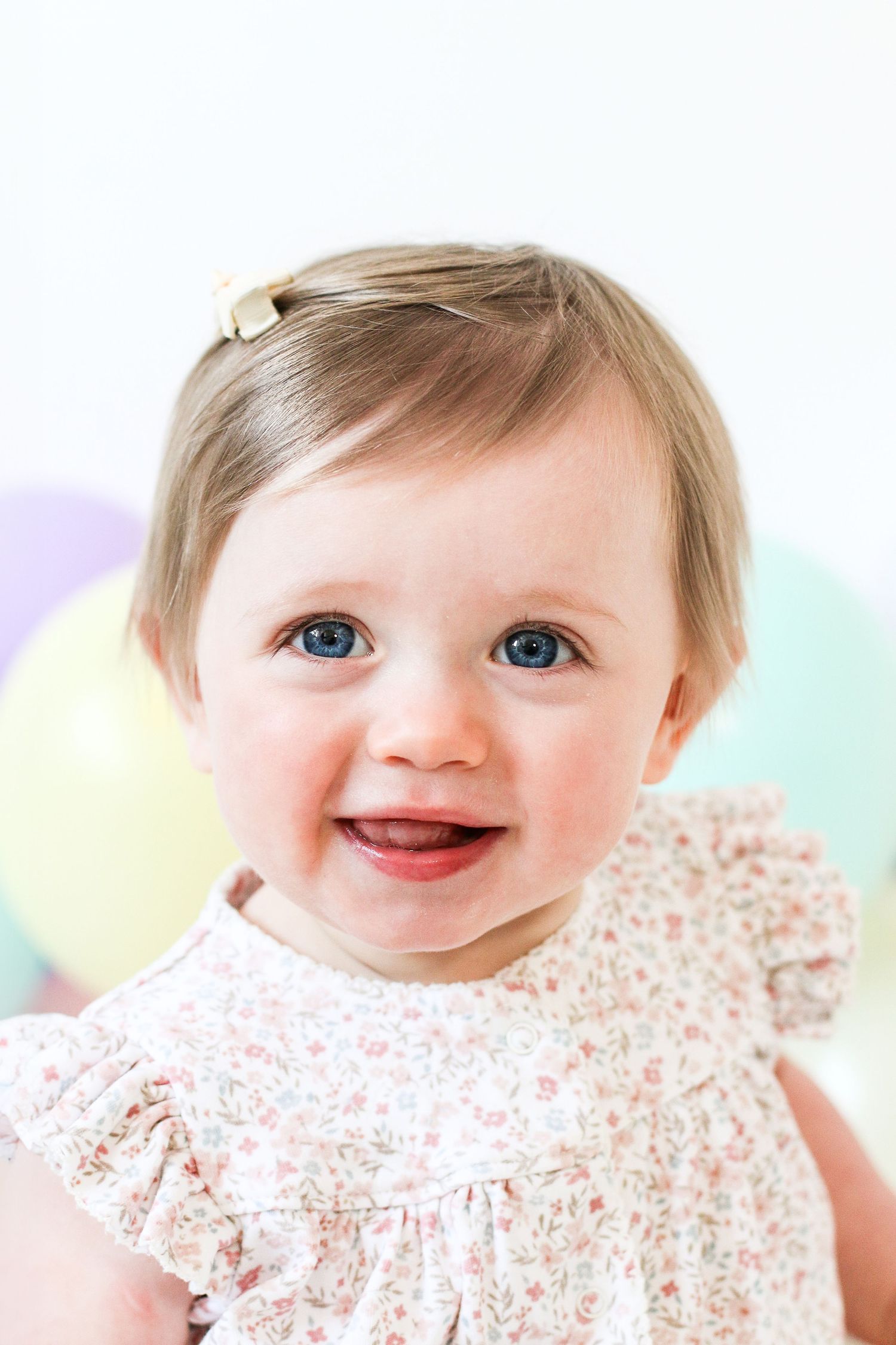Baby wearing floral dress smiles brightly at camera in series of close-up shots with pastel background, during birthday shoot in Nottingham