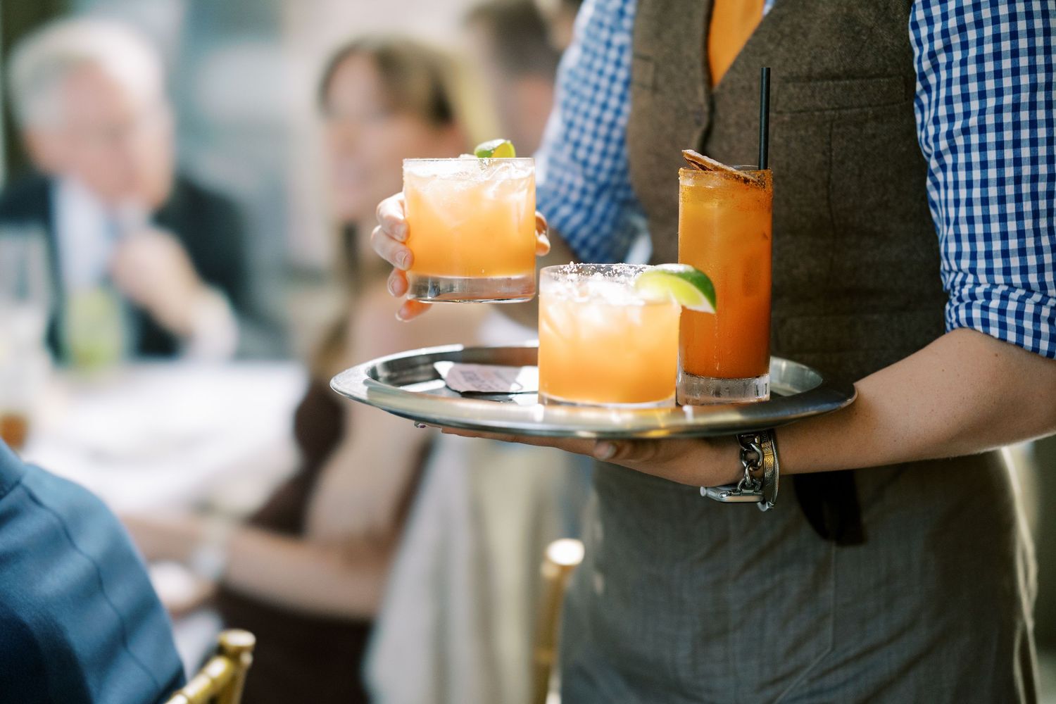 A waiter in a gingham checked shirt and orange vest carries multiple colorful cocktails on a serving tray at Ninety Acres and Pendry Natirar
