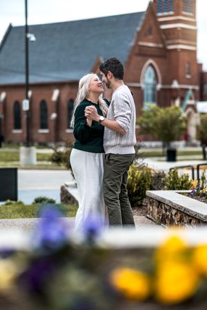 A romantic couple embraces near a brick church with colorful flowers in the foreground on a sunny day.