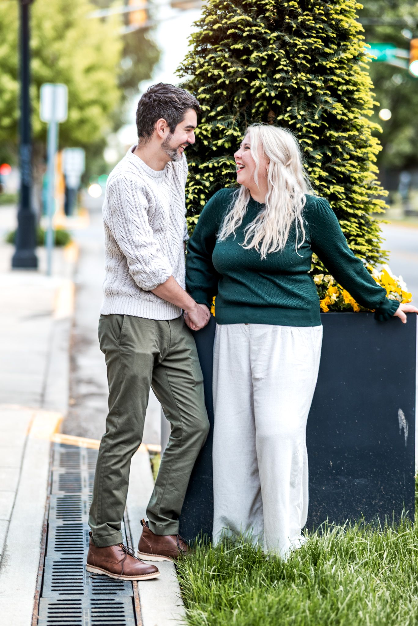 A couple in casual fall attire share a happy moment together on a sidewalk near green hedges and flowers.