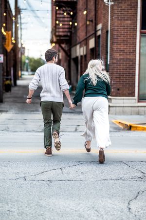 Two people walking down an urban street with brick buildings and string lights, seen from behind in casual attire.