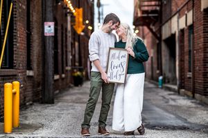 A romantic couple stands close together in an urban alley while holding a 'Best Day Ever' sign between them.