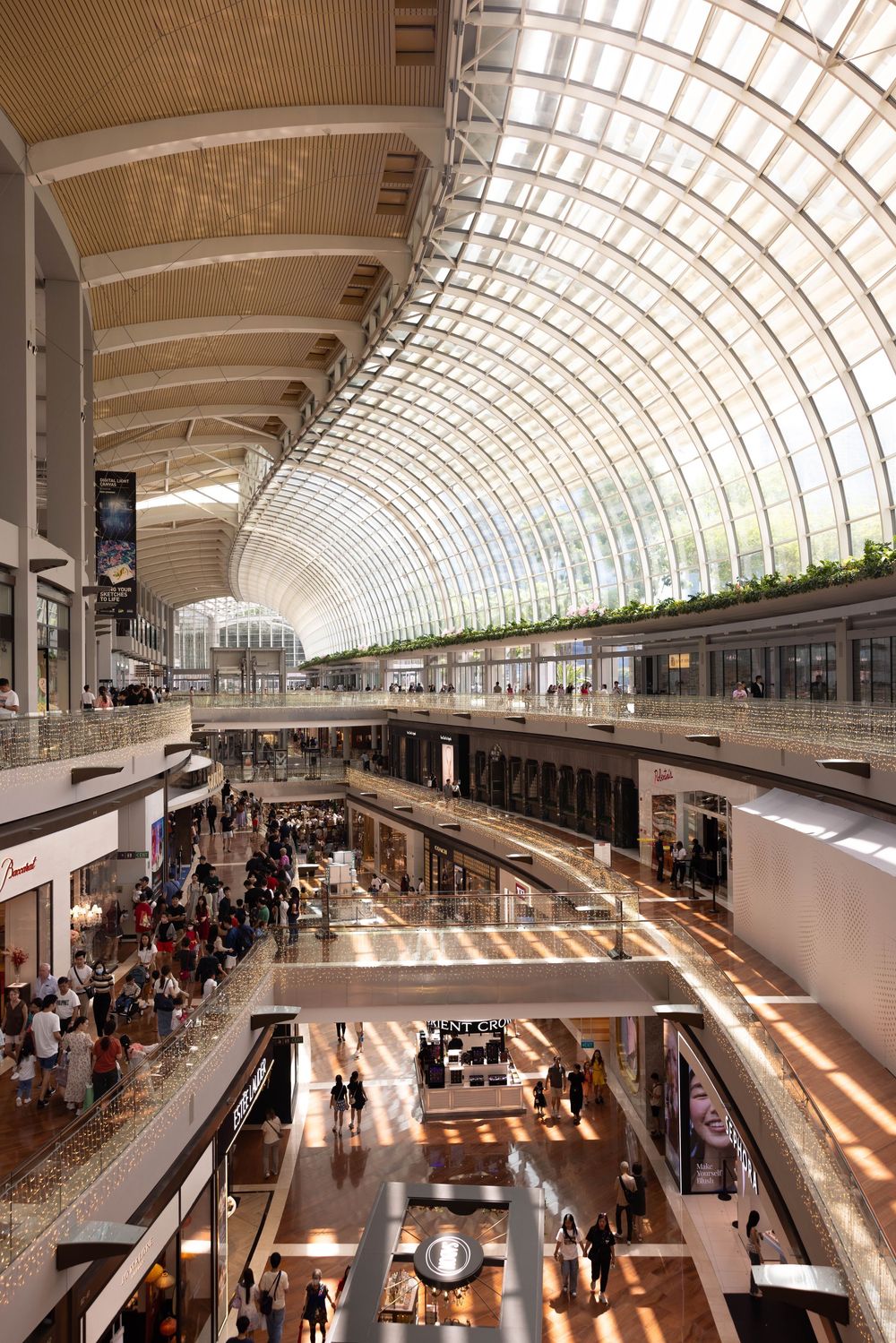 Modern shopping mall interior with curved glass ceiling, multiple retail levels and natural light streaming through atrium design.