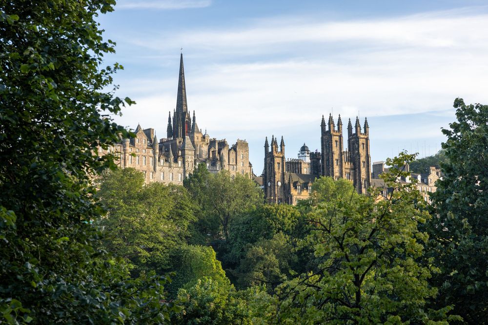 Gothic spires of Edinburgh's historic skyline rise above green treetops against a blue summer sky.