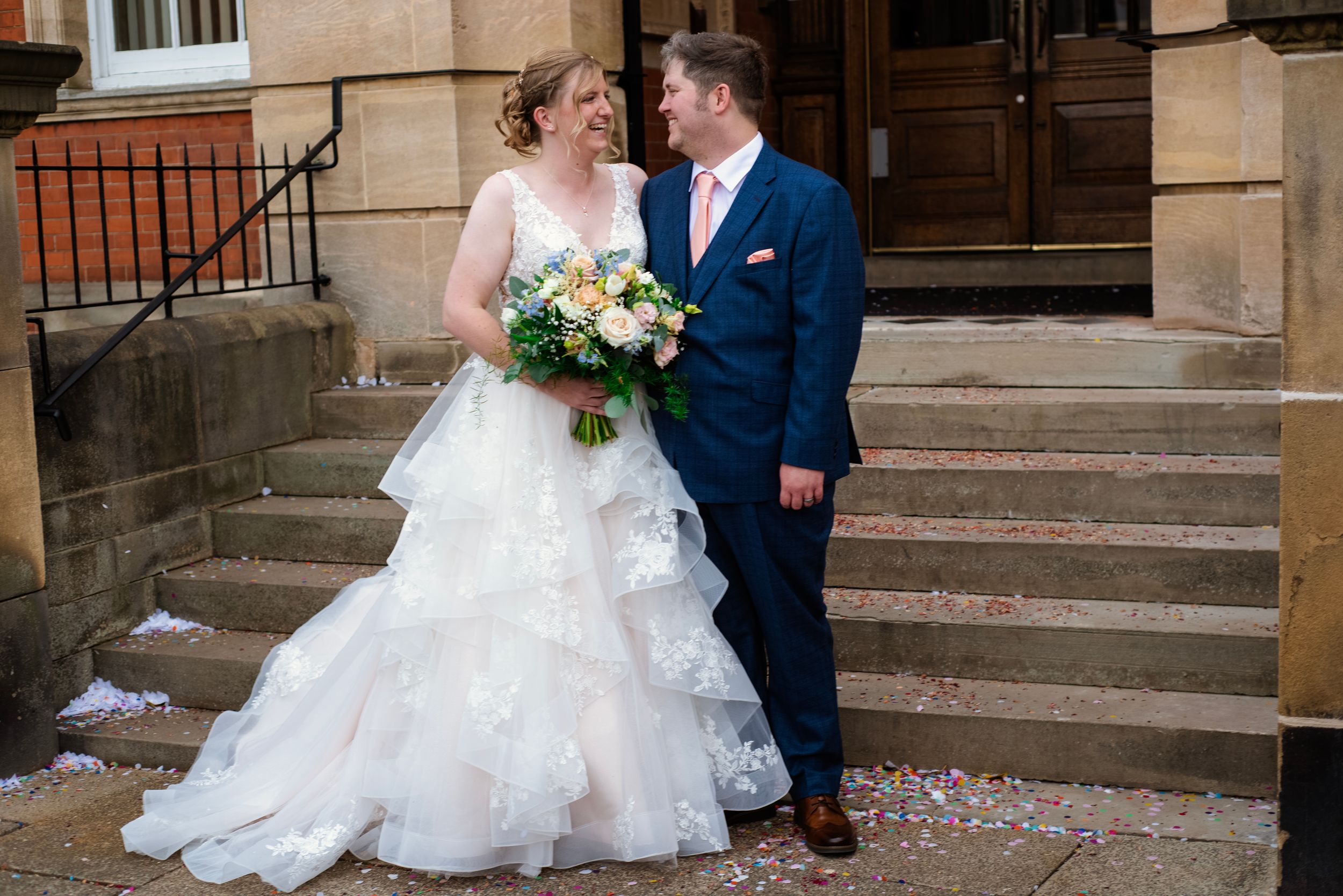 A happy couple stands on stone steps with confetti scattered around, the bride in white and groom in navy blue.