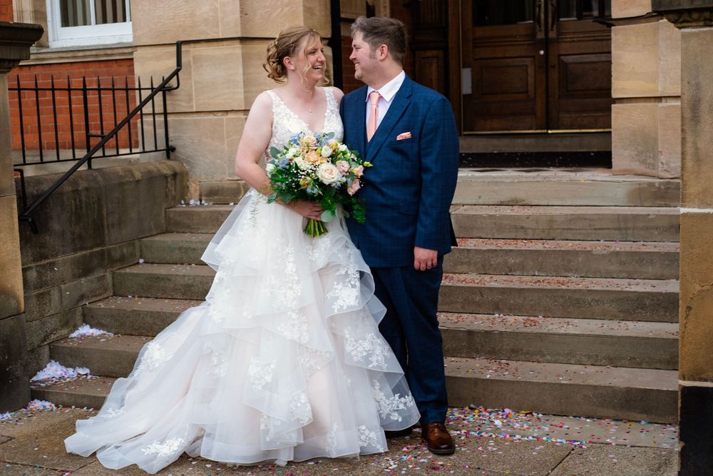 A happy couple stands on stone steps with confetti scattered around, the bride in white and groom in navy blue.