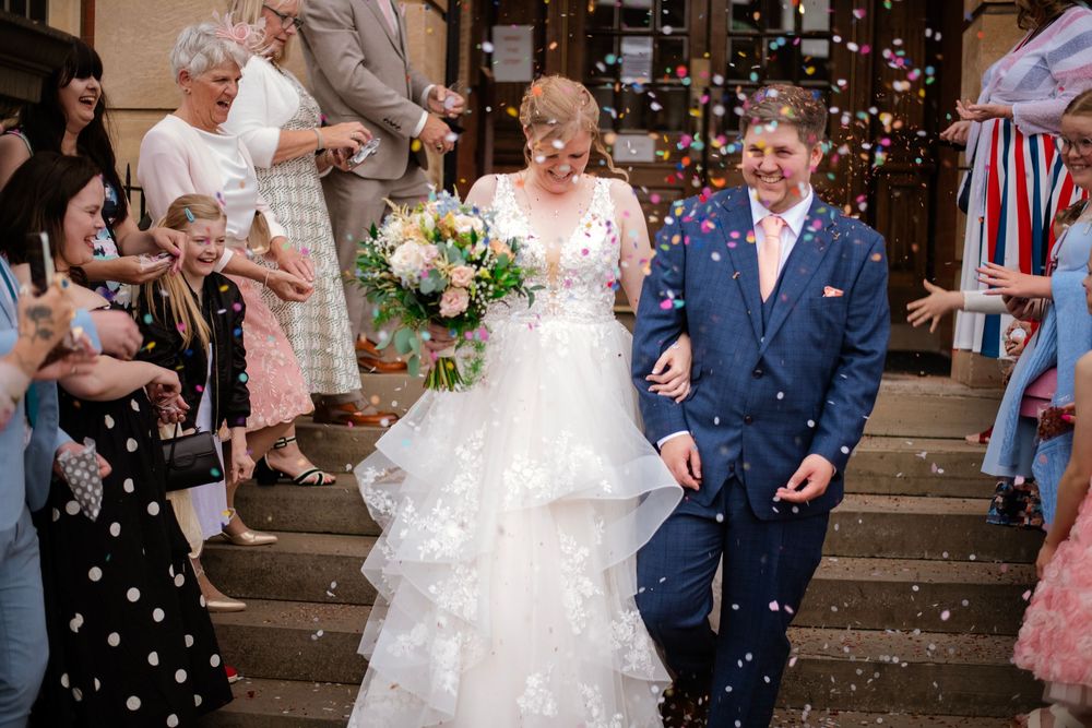 Guests shower wedding party with confetti and bubbles outside a church on a sunny celebration day.