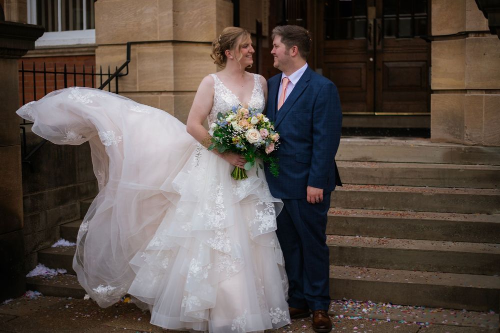 A bride in a flowing white dress with veil holding a colorful bouquet stands with groom in navy suit on stone steps.