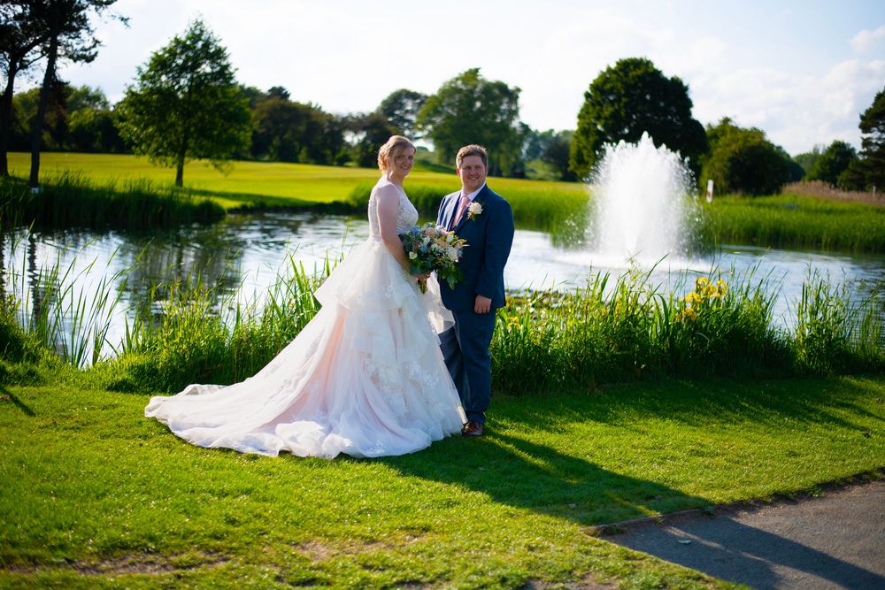 A newlywed couple poses beside a tranquil pond with a fountain and lush green grounds on their wedding day.