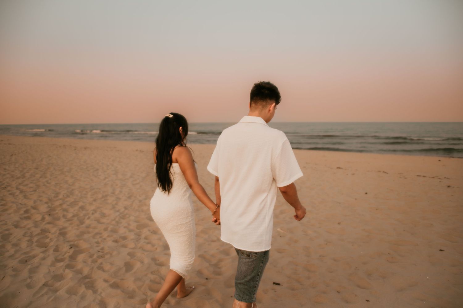 A couple holding hands walks along a sandy beach at sunset with the ocean waves in the background.