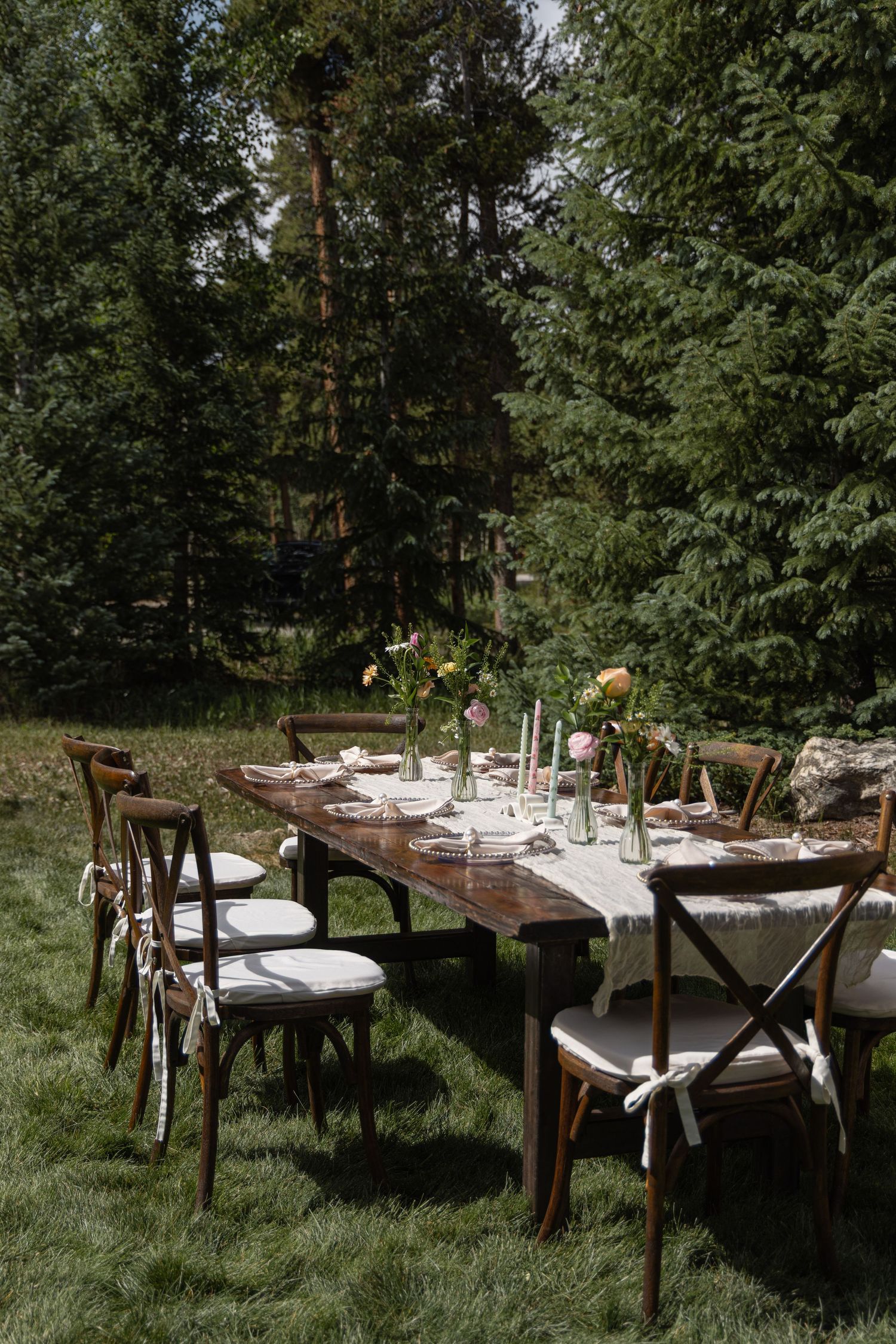Rustic outdoor dining table setup with wooden chairs and white table settings arranged in a garden setting.