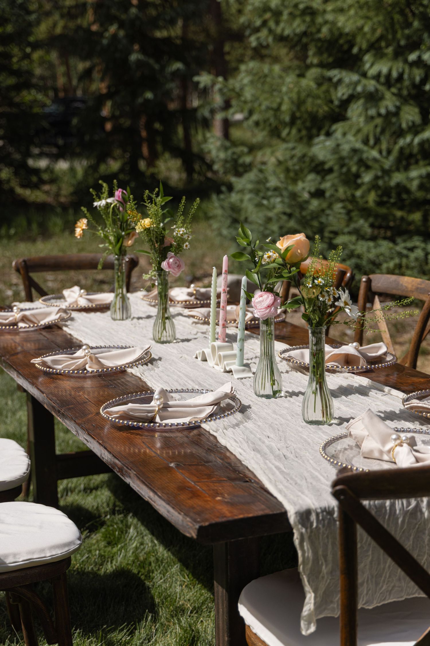 Rustic outdoor dining table settings with delicate floral centerpieces and vintage plates on white linen runners in a garden.