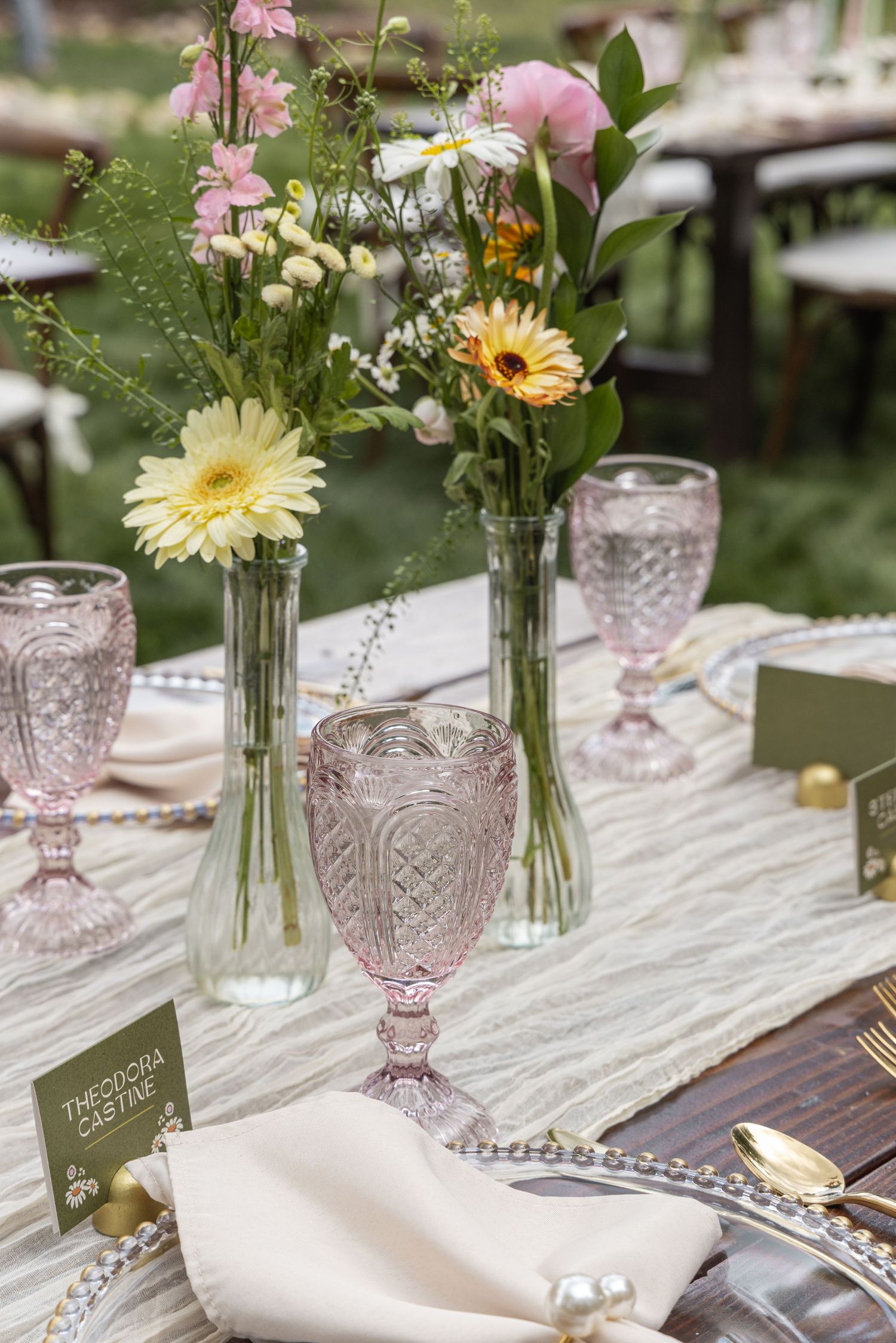 Elegant table setting with pink vintage glassware, white napkins, and fresh pink and yellow flowers in glass vases.
