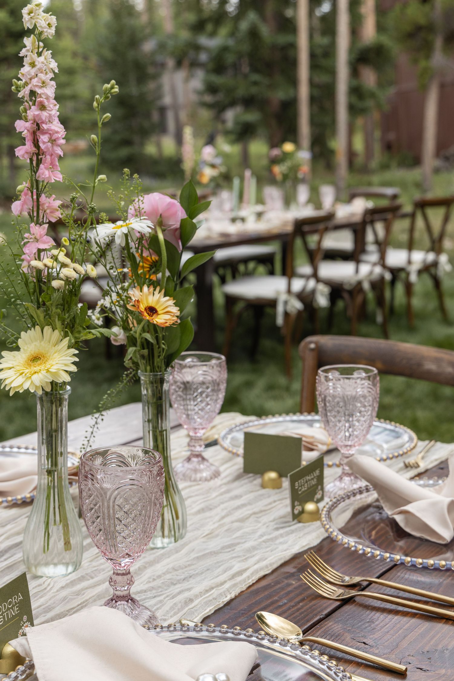 Elegant table setting with pink and yellow flowers in crystal vases and delicate glassware on white linens.