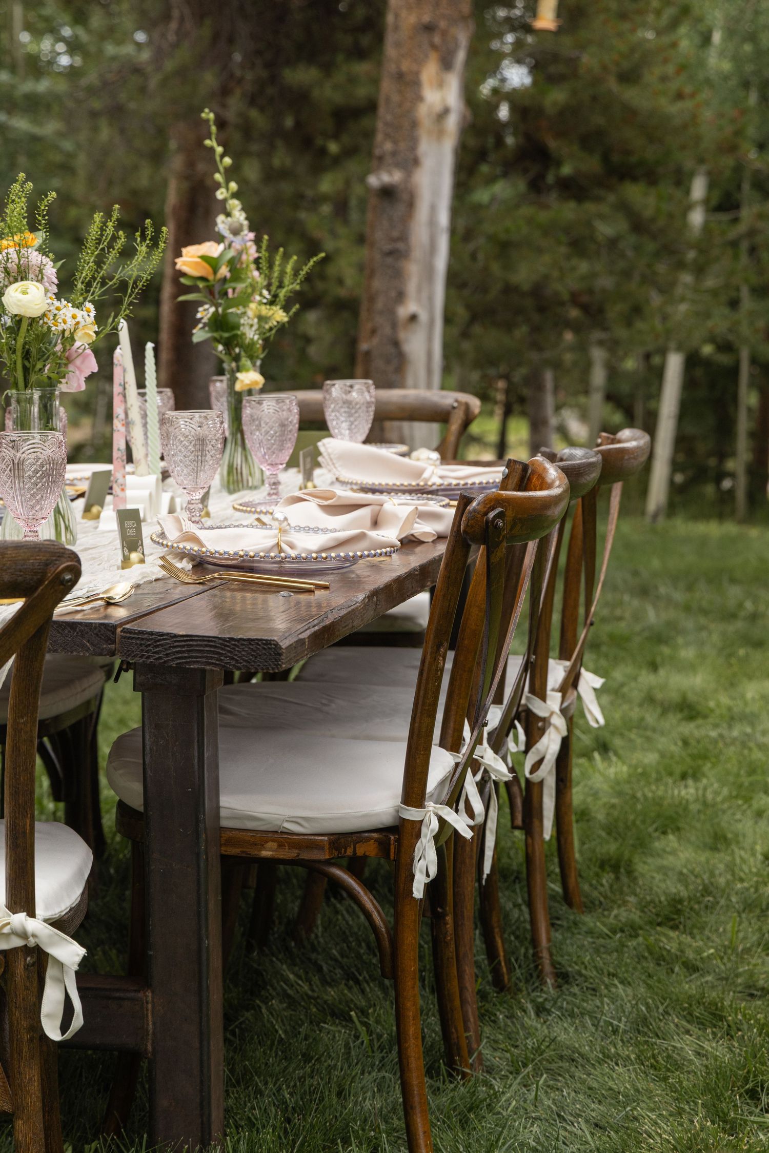 Rustic outdoor dining setup with long wooden tables decorated with delicate glass vases and greenery in a garden setting.