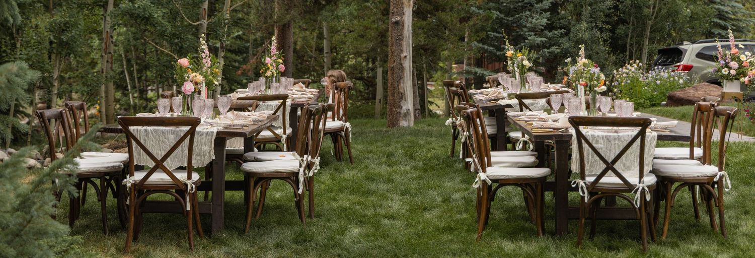 An elegant outdoor garden wedding reception setup with rustic wooden chairs and tables arranged beneath tall pine trees.