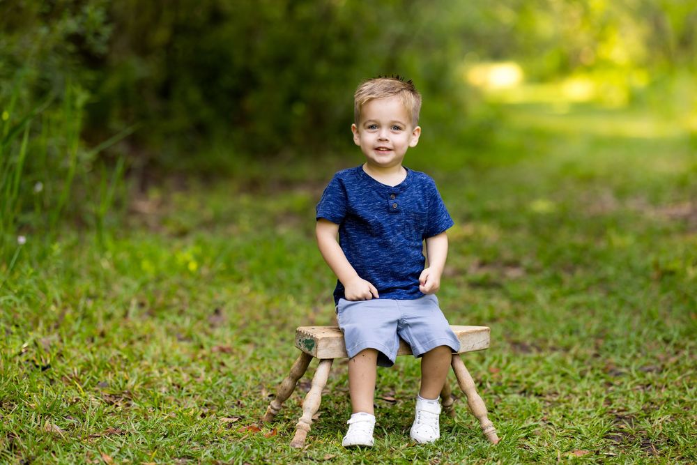 A small child sits on a rustic wooden stool outdoors wearing a navy shirt and grey shorts.