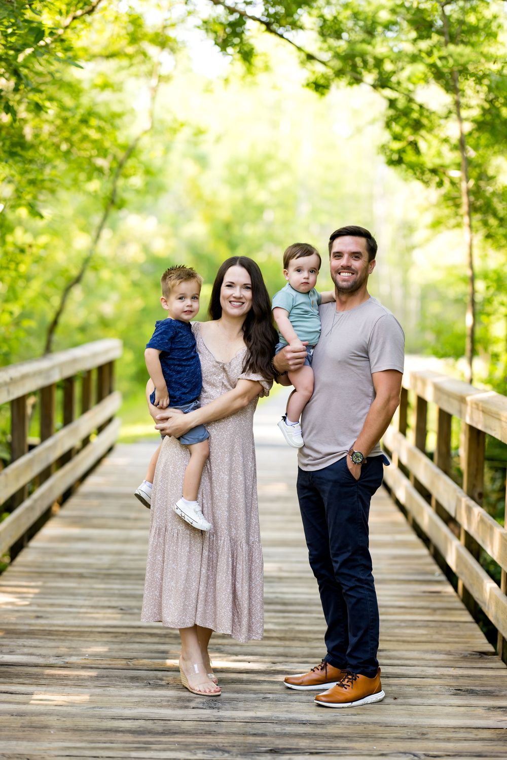 A family stands together on a wooden bridge surrounded by lush green trees on a bright summer day.