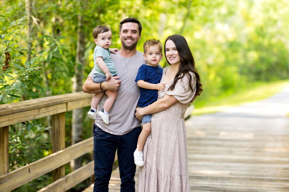 Family group walking and posing together on a wooden bridge pathway surrounded by lush green trees.