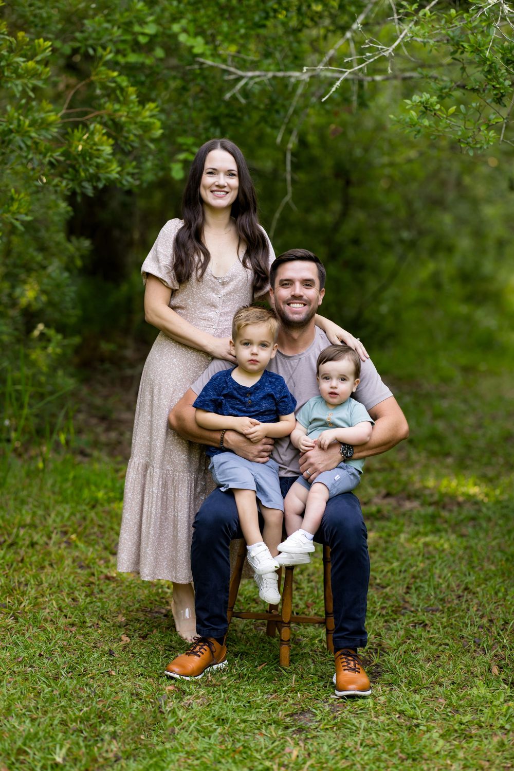 Family wearing casual clothing sits together on grassy ground in a lush green outdoor setting during a photo session.