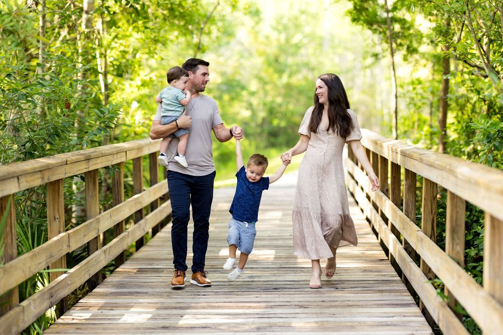 Family walking hand in hand across wooden bridge surrounded by lush green trees in afternoon sunlight.