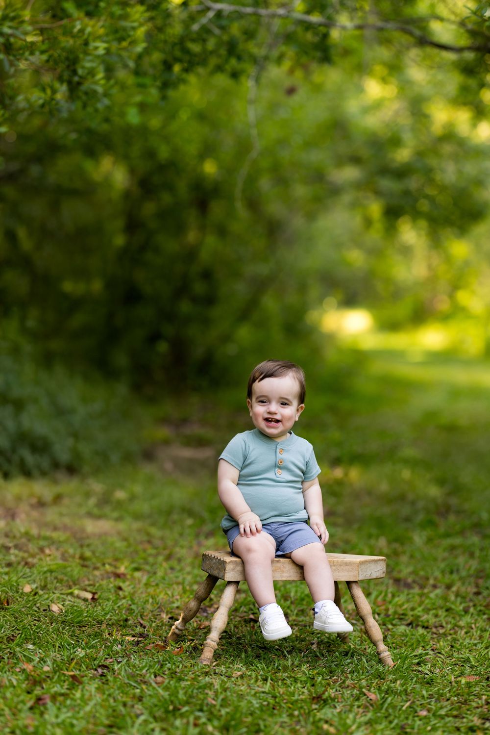 Small child in a light blue outfit sits on a rustic wooden stool in a sunlit garden setting.