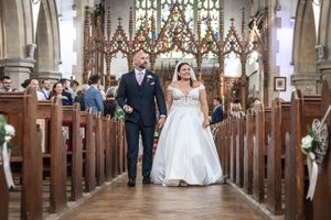 Bride and groom walking down church aisle decorated with beautiful stained glass windows and wooden pews.