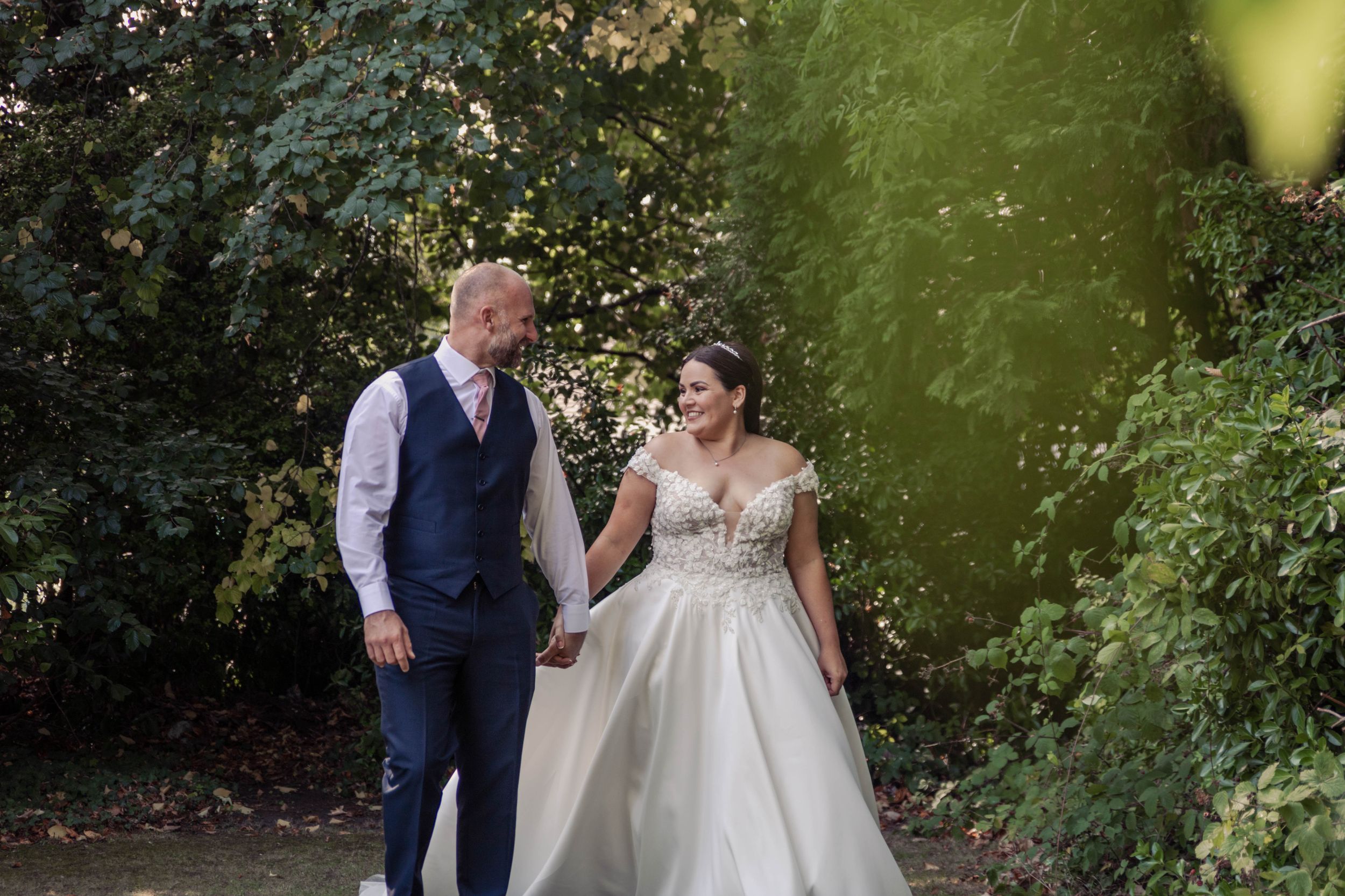 A couple in wedding attire walks together on a garden path surrounded by lush greenery during a summer day.