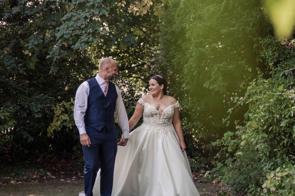 A couple in wedding attire walks together on a garden path surrounded by lush greenery during a summer day.
