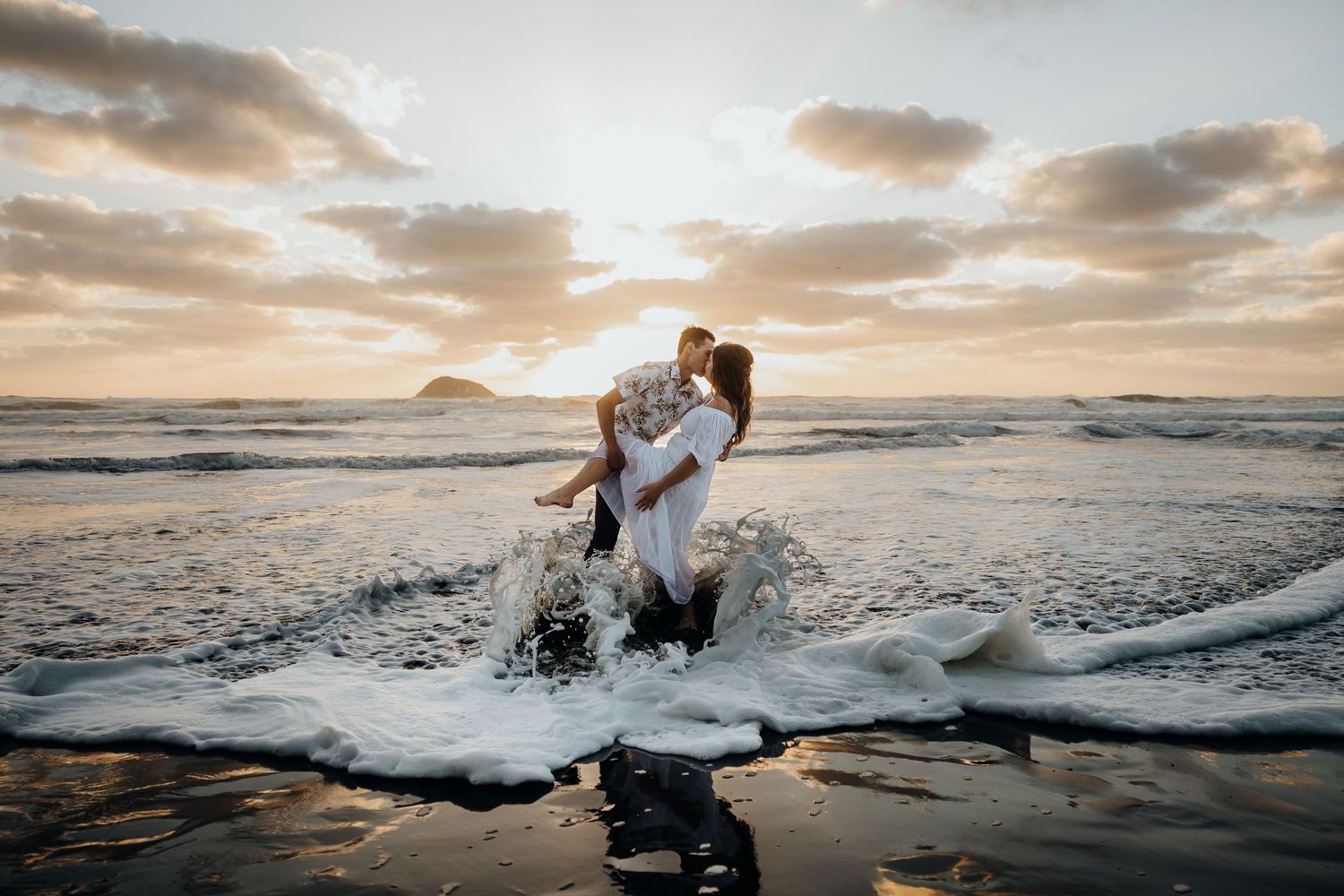 A couple embraces on a beach during golden hour sunset as ocean waves crash around them on the sand.