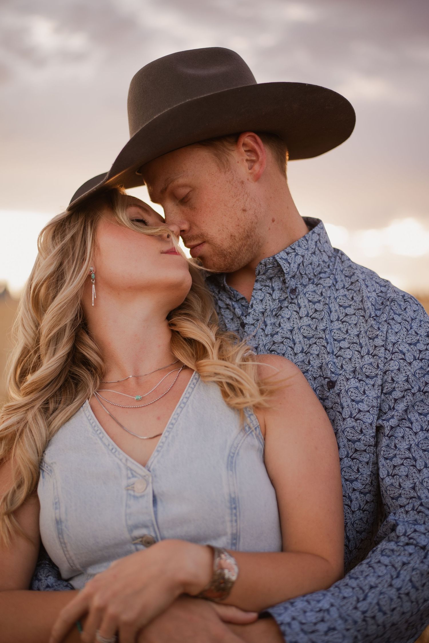 A couple in western attire sharing an intimate moment at sunset in a rural setting.